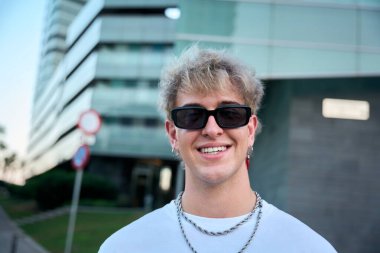 Şık güneş gözlüğü ve gümüş zincirler takarak gülümseyen genç kentsel etkenci, gösterişli modern bir binanın önünde güvenle duruyor, mutluluk ve moda titreşimleri yayıyor.