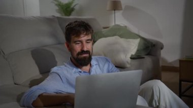 Genç beyaz adam bir kanepede oturuyor ve evdeki dizüstü bilgisayarında çalışıyor, serbest çalışmayı, uzaktan çalışmayı ve dijital girişimciliği temsil ediyor..