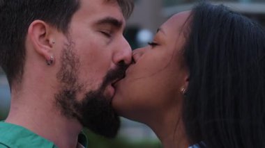 Sevgi dolu ırklar arası bir çift, modern şehir binaları ve yeşillikler tarafından çerçevelenmiş, tutkulu ve yoğun bir açık hava öpücüğünü paylaşır, samimiyet, şefkat ve şehir romantizmini yakalar..