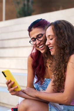 Dikey. İki neşeli genç kadın açık havada oturmuş, akıllı telefondan komik bir içeriğin tadını çıkarıyorlar, neşeli ve bağlı bir an yaratıyorlar.