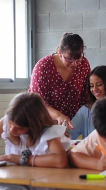 Dikey. İlköğretim öğretmeni öğrencilere okul ödevlerinde yardım ederken, diğer çocuklar da masalarındaki not defterlerine yazmaya odaklanırken genç bir çocuğa rehberlik ediyor.