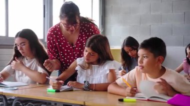 İlköğretim öğretmeni öğrencilere okul ödevlerinde yardım ederken, diğer çocuklar da masalarındaki not defterlerine yazmaya odaklanırken genç bir çocuğa rehberlik ediyor.