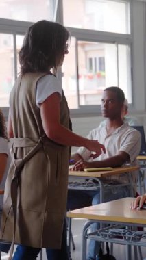 Dikey. Bir bayan öğretmen sınıfta sıraların arasından yürür ve şüpheleri gidermek, ders sırasında etkileşimi, rehberliği ve akademik desteği teşvik etmek için öğrenciye yaklaşır..