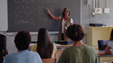 Bir öğretmen tahtadaki dersi, öğrencilerin arkasındaki sahnenin çerçevelendiği notlara işaret ederek anlatıyor, talimatlar, katılım ve odaklanmış sınıf öğrenimi vurguluyor..