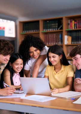 Dikey. Beş farklı lise öğrencisi gülümsüyor ve kütüphanede bir proje üzerinde birlikte çalışıyorlar, dizüstü bilgisayar kullanıyorlar ve notlar alıyorlar, takım çalışmasını ve eğitimdeki işbirliğini gösteriyorlar