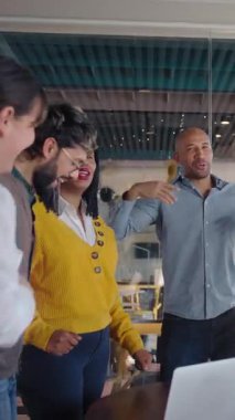 Dikey. Çok kültürlü meslektaşlar, bir masanın etrafında toplanmış, parlak, modern bir ofiste, birlik, motivasyon ve işbirlikçi ruhu ifade eden iyi haberlerin ardından gülümsüyor ve tezahürat yapıyorlar..