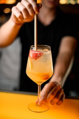 Buzlu, limonlu ve ahududu dilimli bir bardak köpüklü kokteyle odaklan. Hangi erkek barmenin eli nazikçe pipetle karıştırır?