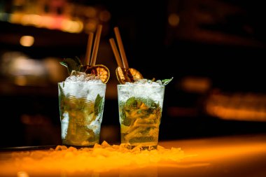 Portakal dilimiyle süslenmiş soğuk mojito kokteyli ve koyu bir barda pipet içilen yakın plan bardaklar.