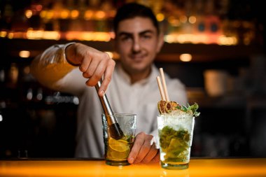 Barın tezgahında mojito kokteyli olan yakın plan bardaklar ve çelik muddler kullanan erkek barmen.