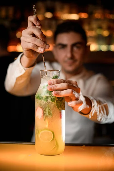 Professional bartender stirring cocktail in bottle with long bar spoon on the bar counter on the blurred background