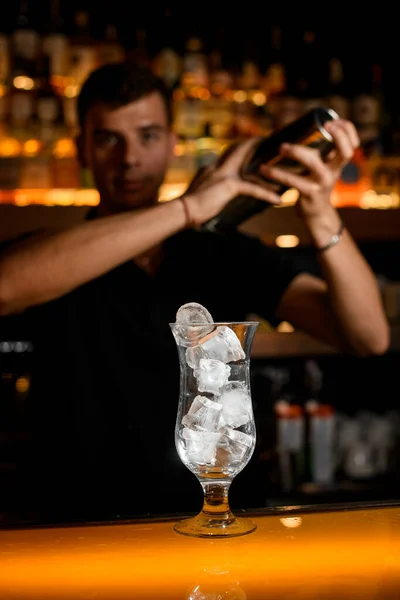 transparent glass with ice cubes on the bar counter. Blurred male bartender with shaker in his hands on background