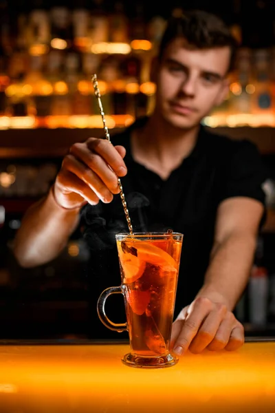 hand of male bartender neatly holds spoon and stirs hot drink with lemon and berries in transparent cup on the bar counter