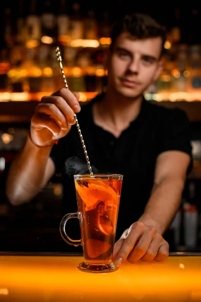 hand of male bartender holds spoon and accurate stirs hot drink with lemon and berries in transparent cup on the bar counter