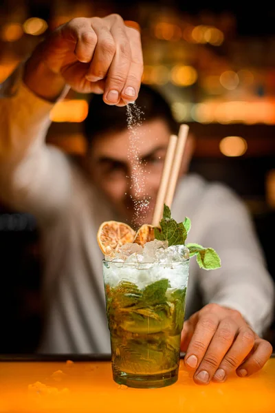selective focus on glass of cold cocktail with mint leaves and ice in which male bartender pours powdered sugar