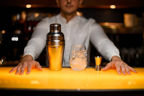stock image steel shaker and glass with ice cubes and jigger and hands of male bartender on yellow bar surface
