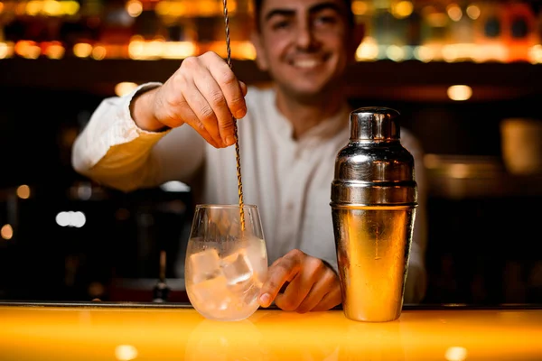 steel shaker and hand of bartender holding long bar spoon and stirring alcoholic cocktail with ice in transparent glass on yellow bar