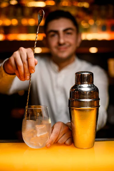close-up on steel shaker and hand of bartender holding long bar spoon and stirring alcoholic cocktail with ice in transparent glass on yellow bar