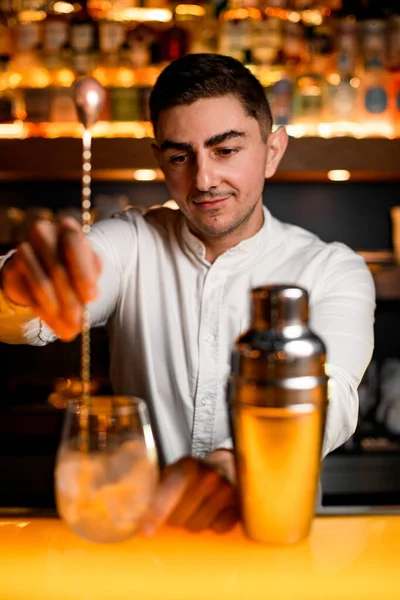 Selective focus of bartender holding long bar spoon and stirring alcoholic cocktail with ice in transparent glass on yellow bar surface