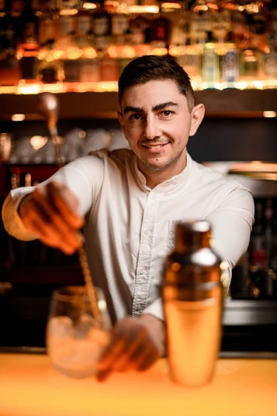 selective focus on young caucasian male bartender mixing cocktail in glass with long bar spoon. Portrait of male barman at bar