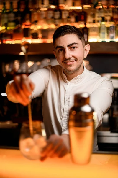 selective focus on face of young caucasian male bartender mixing cocktail in glass with long bar spoon. Portrait of male barman at bar