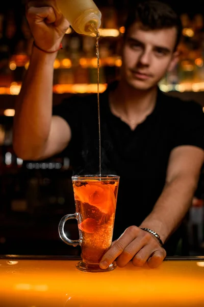 male masterfully bartender adds syrup to tea drink with lemon slices in transparent cup on the bar counter