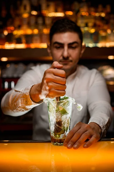 Erkek barmenin eli bardağı karıştırıyor ve şeffaf bardakta mojito malzemelerini ustaca sıkıyor.