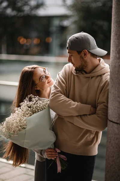 白い花の花束を手にした若い笑顔の女性はキャップを持つ男を見ています 恋のカップル ラブストーリー — ストック写真
