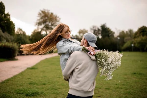 若い男は花の花束で美しい女の子を持ち上げ 公園で彼女を旋回させます ラブストーリー — ストック写真