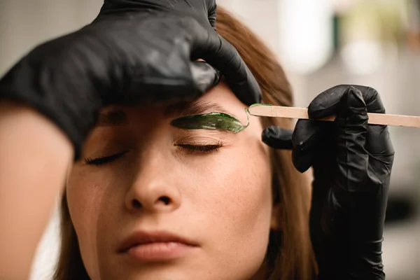 Process of removing hair eyebrows with special green wax on the skin of woman. Professional correction in beauty salon.