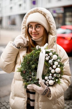 Şapka ceketli ve eldivenli genç bir kadın beyaz toplar ve kurdelelerle süslenmiş ladin ve thuja dallarından oluşan bir Noel çelengi tutuyor.