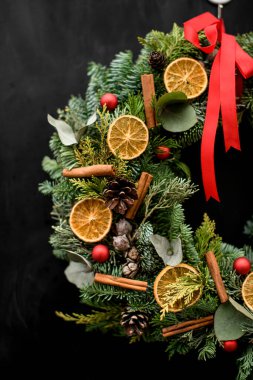 Kuru portakal dilimleri, tarçın topları ve koyu arkaplanda kurdeleyle süslenmiş yuvarlak parlak noel çelenginin kesilmiş fotoğrafı.