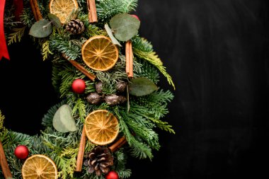 Kuru portakal dilimleri, tarçın topları ve koyu arkaplanda kurdeleyle süslenmiş yuvarlak parlak noel çelenginin bir kısmı.