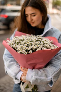 Güzel, yemyeşil bir buket çiçek, küçük kasımpatılar ve papatyalar genç ve çekici bir kadının ellerinde pembe ambalaj kağıdında.