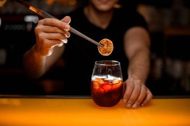 Şeffaf cam, soğuk alkollü içecek ve erkek eli üzerinde cımbızla kuru portakal dilimi tutuyor.