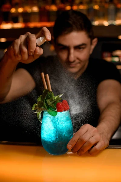 man bartender masterfully sprinkles on glass of cold blue cocktail decorated with mint and red raspberries. Signature cocktail