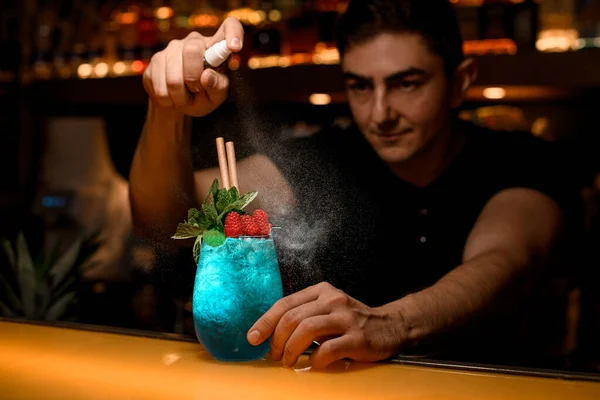 man bartender skillfully sprinkles on glass of cold blue cocktail decorated with mint and red raspberries. Signature cocktail