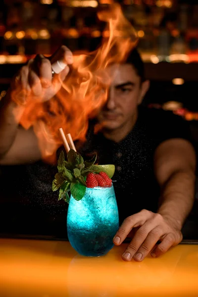 man bartender sprinkles and set fire glass of cold blue cocktail decorated with mint and red raspberries. Signature cocktail