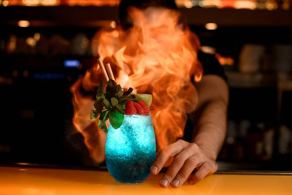 man bartender gently sprinkles and set fire glass of cold blue cocktail decorated with mint and red raspberries. Signature cocktail