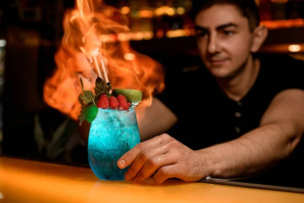 man bartender masterfully sprinkles and set fire glass of cold blue cocktail decorated with mint and red raspberries. Signature cocktail