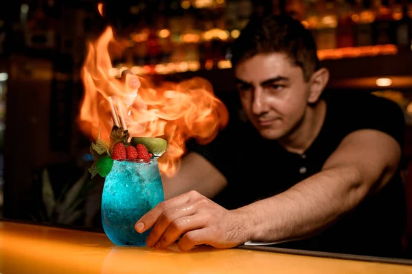 man bartender skillfully sprinkles and set fire glass of cold blue cocktail decorated with mint and red raspberries. Signature cocktail