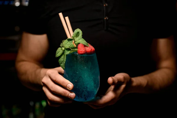 hands of man bartender accurate holds glass with blue cocktail decorated with mint and red raspberries. Cropped photo. Signature cocktail. Close-up