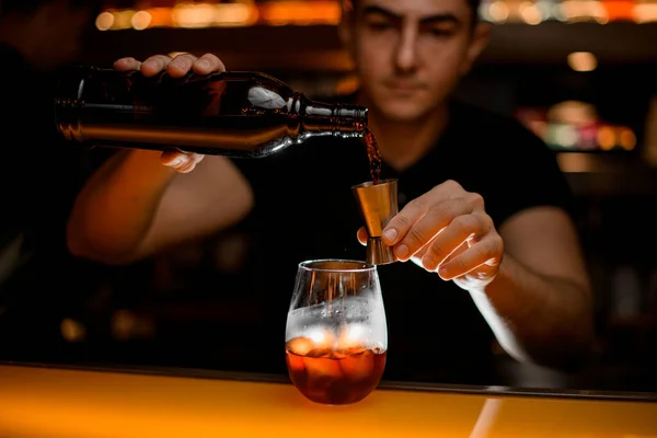 Close-up of bottle and jigger in hands of male bartender and glass with iced drink on bar. Process of making cocktail.
