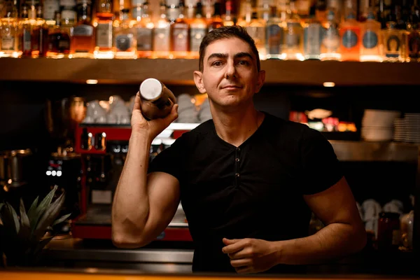 Handsome male barman in bar interior shaking and mixing alcohol cocktail. Professional bartender at work in night club with shaker in hands.