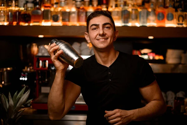 Smiling male barman in bar interior shaking and mixing alcohol cocktail. Professional bartender at work in night club with shaker in hands.