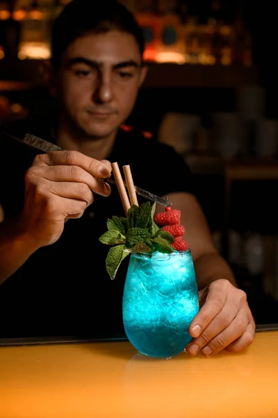 man bartender accurate decorate glass with bright blue cold cocktail with mint by red raspberries. Signature cocktail