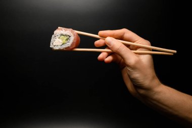 great side view of Philadelphia roll in hand holding with bamboo chopsticks on dark background. One piece of roll Philadelphia with salmon and cream cheese.