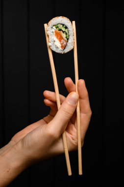 side view of sushi roll california with salmon, cream cheese, cucumber, sesame seeds in hand holding with bamboo chopsticks on dark background. Japanese oriental cuisine. Sushi menu.