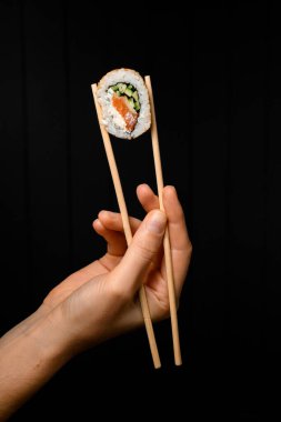side view of one sushi roll california with salmon, cream cheese, cucumber, sesame seeds in hand holding with bamboo chopsticks on dark background. Japanese oriental cuisine. Sushi menu.