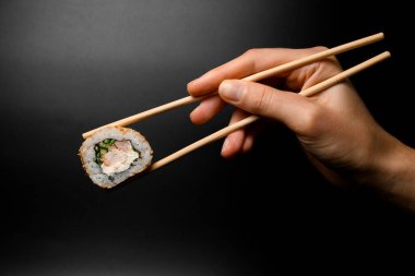 hand holding one piece of roll california with snow crab meat, cream cheese, cucumber garnished sesame seeds with bamboo chopsticks on dark background.