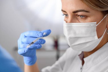 close-up of the face of female doctor in protective medical mask with blue latex gloves on her hands who holds a syringe with liquid